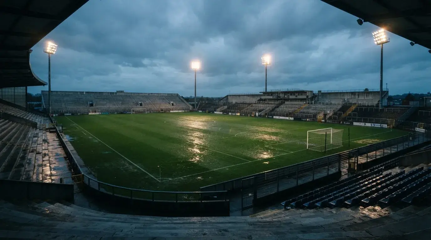 Ein Fußballplatz mit Flutlichtmasten in der Abenddämmerung, aus der Ferne gesehen, leere Tribünen und nasser Rasen unter bewölktem Himmel.