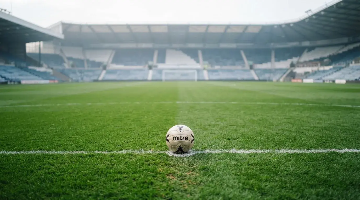 Ein Fußball liegt auf dem Elfmeterpunkt eines leeren Rasenplatzes, im Hintergrund eine Tribüne im Dunst.