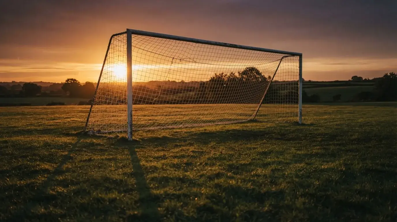 Ein Fußballtor mit Netz bei Sonnenuntergang auf einem leeren Platz, die Schatten der Torpfosten liegen lang auf dem Gras.