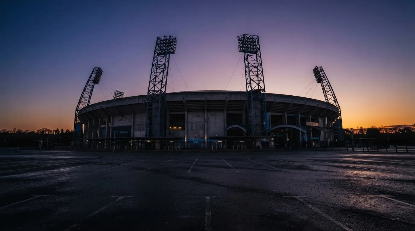Ein Fußballstadion von außen in der Dämmerung, hohe Flutlichtmasten vor einem violetten Himmel, davor ein leerer Parkplatz.