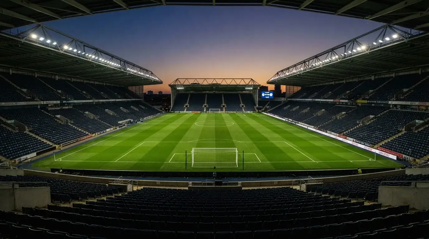 Eine leere Stadiontribüne unter Flutlicht aus der Ferne betrachtet, die Rasenfläche leuchtet grün in der Dämmerung.