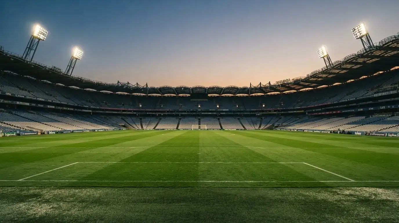 Eine leere Stadiontribüne unter Flutlicht aus der Ferne betrachtet, die Rasenfläche leuchtet grün in der Dämmerung.