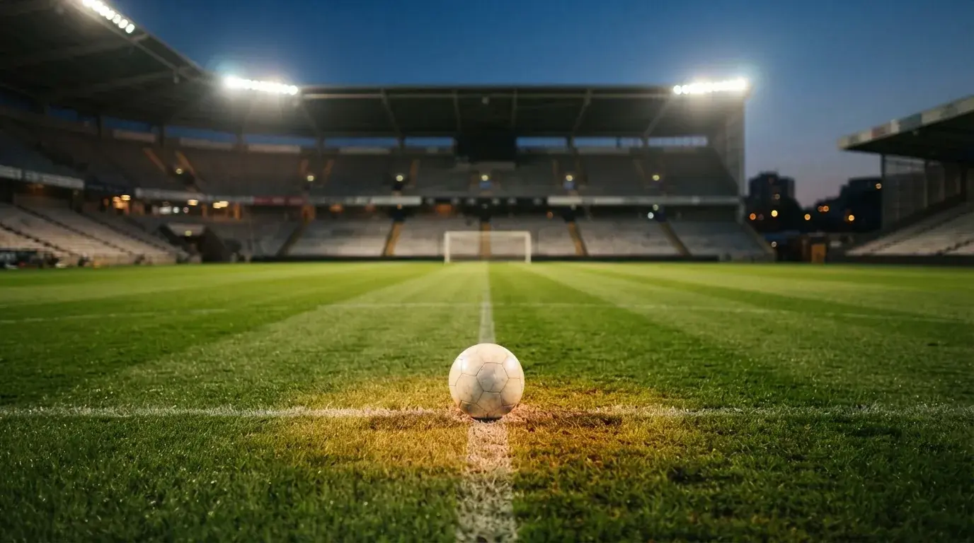 Ein Fußball liegt auf dem Rasen eines leeren Platzes, im Hintergrund ein Stadion unter Flutlicht in der Abenddämmerung.