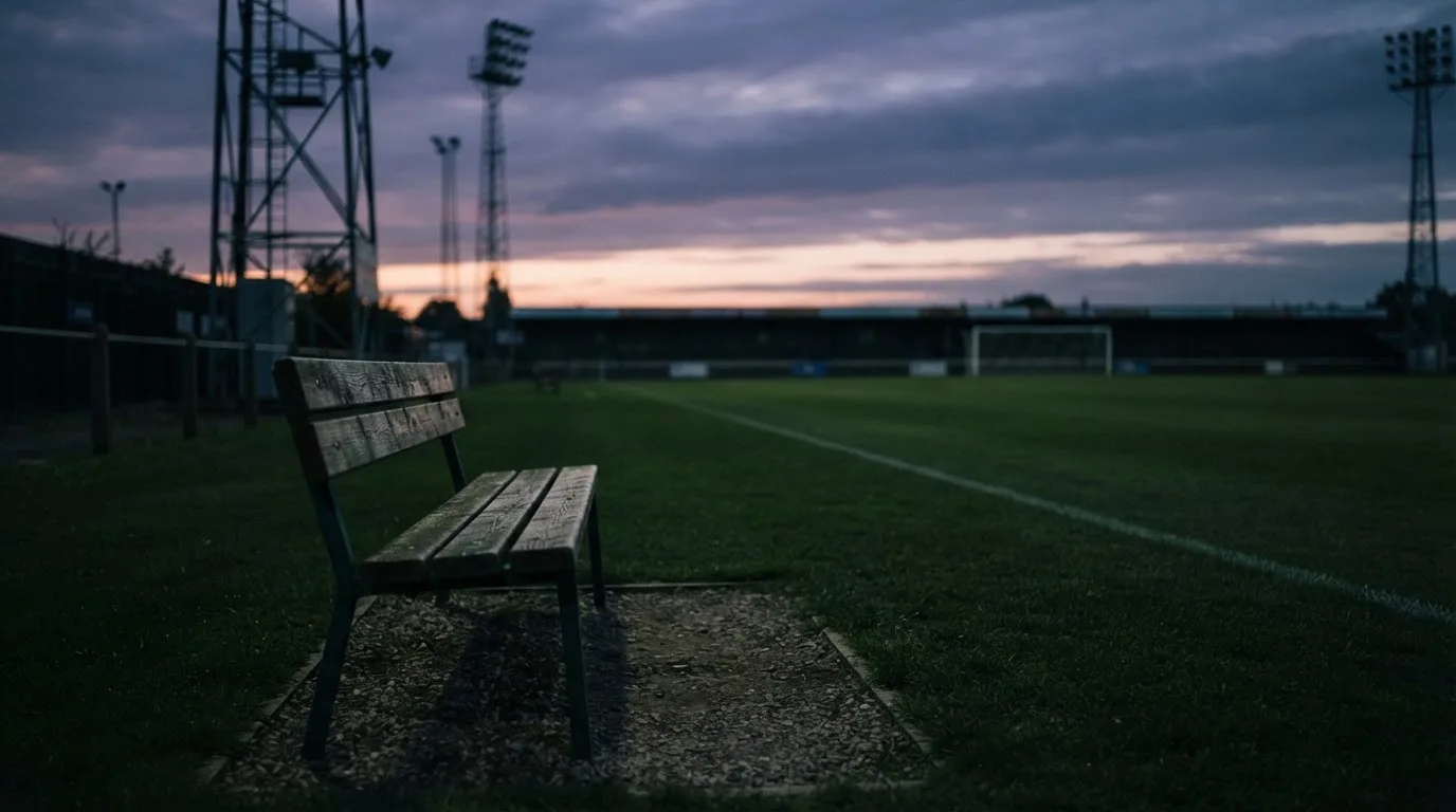 Eine leere Parkbank am Rand eines Fußballfelds in der Abenddämmerung, im Hintergrund ragen dunkle Flutlichtmasten in den Himmel.