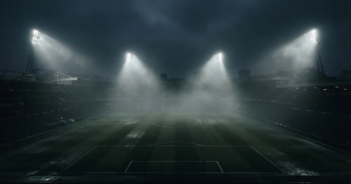 Ein Stadion unter Flutlicht, aus großer Entfernung über eine dunkle Wiese fotografiert, am Himmel zieht Nebel auf.